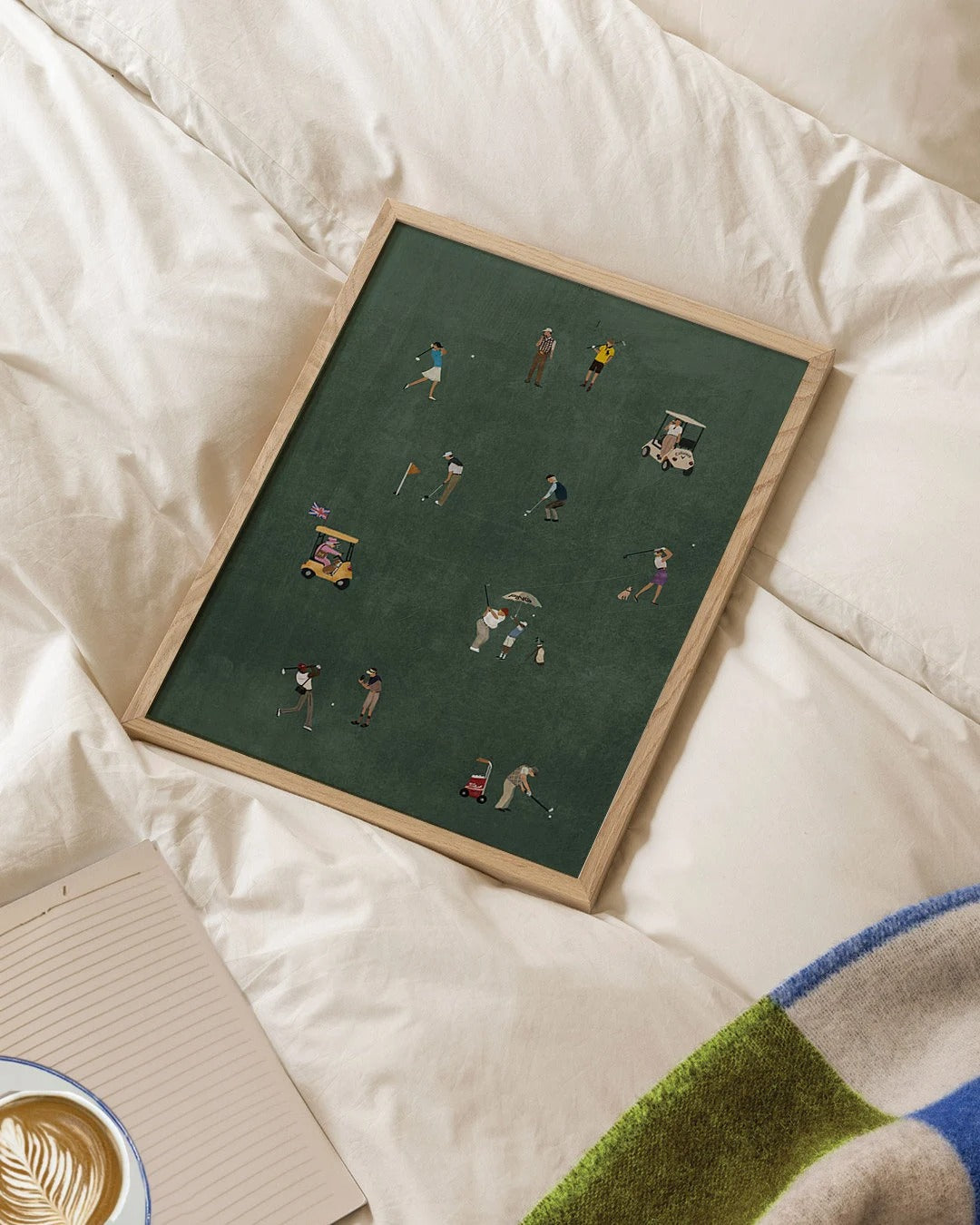 Framed artwork of a golf scene on a bed with a cup of coffee and notebook.