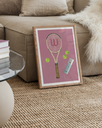 Pink tennis racket art print in a wood frame beside a beige sofa.